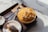 A delicious-looking dessert featuring a bowl of mango shaved ice topped with chunks of cheesecake sits on a dark serving tray. Next to it, a small plate holds an almond croissant. Several silver spoons rest on a beige napkin beside the bowl, all placed on a marble surface.