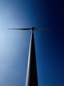 A wind turbine is viewed from below, silhouetted against a clear blue sky. The tall, slender tower extends upwards, culminating in three blades that are aligned horizontally.