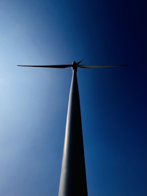 A wind turbine is viewed from below, silhouetted against a clear blue sky. The tall, slender tower extends upwards, culminating in three blades that are aligned horizontally.