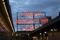 A large, illuminated neon sign reads 'Public Market Center' with a round clock to its side. The setting appears to be in the evening or early night, with a clear view of the vibrant red signage against the darkening blue sky. Below the sign, there are market stalls or storefronts with visible lighting.