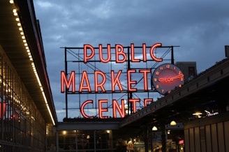 A large, illuminated neon sign reads 'Public Market Center' with a round clock to its side. The setting appears to be in the evening or early night, with a clear view of the vibrant red signage against the darkening blue sky. Below the sign, there are market stalls or storefronts with visible lighting.