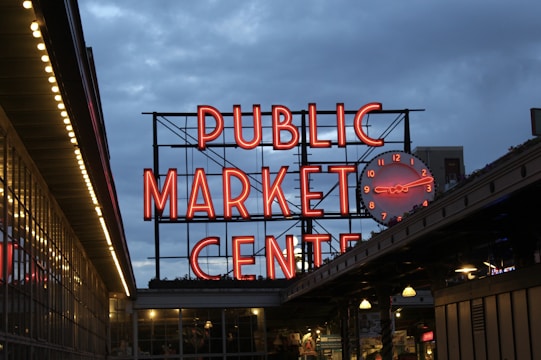 A large, illuminated neon sign reads 'Public Market Center' with a round clock to its side. The setting appears to be in the evening or early night, with a clear view of the vibrant red signage against the darkening blue sky. Below the sign, there are market stalls or storefronts with visible lighting.