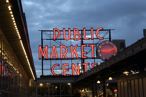 A large, illuminated neon sign reads 'Public Market Center' with a round clock to its side. The setting appears to be in the evening or early night, with a clear view of the vibrant red signage against the darkening blue sky. Below the sign, there are market stalls or storefronts with visible lighting.