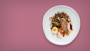 A white plate containing a variety of foods including a piece of glazed salmon garnished with sesame seeds, a half-cut boiled egg, and a colorful mix of diced vegetables on a pink background.