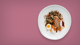 A white plate containing a variety of foods including a piece of glazed salmon garnished with sesame seeds, a half-cut boiled egg, and a colorful mix of diced vegetables on a pink background.