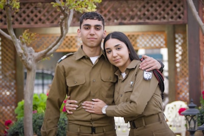 A young male and female, both dressed in matching brown military uniforms, are standing closely together outdoors. The woman has her arm around the man's waist, and there are trees and lattice structures in the background, suggesting a garden setting.
