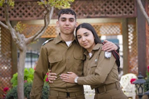 A young male and female, both dressed in matching brown military uniforms, are standing closely together outdoors. The woman has her arm around the man's waist, and there are trees and lattice structures in the background, suggesting a garden setting.