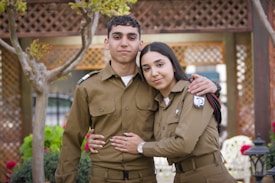 A young male and female, both dressed in matching brown military uniforms, are standing closely together outdoors. The woman has her arm around the man's waist, and there are trees and lattice structures in the background, suggesting a garden setting.