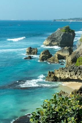 A scenic coastal view featuring turquoise water and rocky cliffs. The waves crash against large boulders scattered along the shoreline. Small plants and greenery are visible in the foreground, adding a touch of lushness to the rugged landscape. In the distance, a stretch of sandy beach and a few small boats can be seen, all under a clear blue sky.