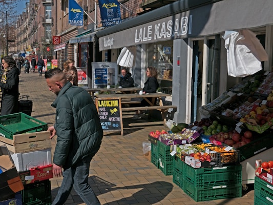 A street scene featuring several people engaged in various activities. On the left, a person is carrying a crate in front of a row of buildings, while on the right, several crates filled with fresh fruits and vegetables are displayed outside a small shop. People are sitting at a table under the shop's awning, enjoying their time. Signs and banners for different businesses, including a tailor and a stone-cleaning service, are visible.