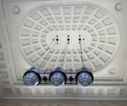 An ornate ceiling featuring a geometric pattern with recessed panels and intricate molding. Four large, decorative pendant lights hang down, each with a metal frame and glass panels, suspended by long chains.
