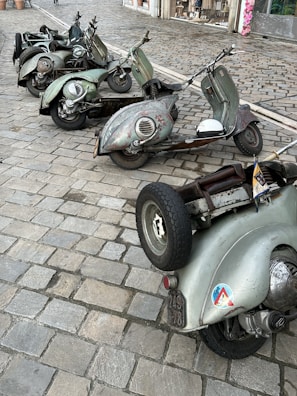 A group of vintage scooters is parked on a cobblestone street. The scooters have a weathered, rustic look with greenish hues and visible wear. In the background, a building with flower pots can be seen, adding a quaint touch to the setting.