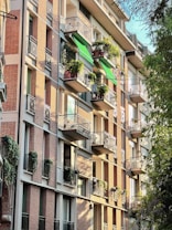 A multi-story apartment building with brick and cream-colored walls features several balconies. Each balcony is adorned with green plants, and some have green awnings. Sunlight casts shadows, creating a warm and inviting atmosphere. Mature trees are present in the foreground.