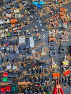 A variety of electrical components are arranged neatly on a dark surface. The components include switches, plugs, connectors, and other hardware, each displaying different colors and forms. The words on the surface, partially visible, seem to be part of a packaging or material brand. An array of colors, shapes, and metal finishes make up the intricate display.