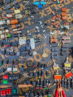 A variety of electrical components are arranged neatly on a dark surface. The components include switches, plugs, connectors, and other hardware, each displaying different colors and forms. The words on the surface, partially visible, seem to be part of a packaging or material brand. An array of colors, shapes, and metal finishes make up the intricate display.