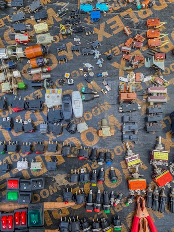 A variety of electrical components are arranged neatly on a dark surface. The components include switches, plugs, connectors, and other hardware, each displaying different colors and forms. The words on the surface, partially visible, seem to be part of a packaging or material brand. An array of colors, shapes, and metal finishes make up the intricate display.