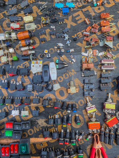 A variety of electrical components are arranged neatly on a dark surface. The components include switches, plugs, connectors, and other hardware, each displaying different colors and forms. The words on the surface, partially visible, seem to be part of a packaging or material brand. An array of colors, shapes, and metal finishes make up the intricate display.