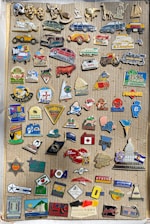 A collection of various vintage pins is displayed on a corrugated cardboard backdrop. The pins feature a variety of designs including cars, animals, flags, logos, and symbols. The pins are colorful and arranged in a somewhat random order, covering most of the cardboard surface.