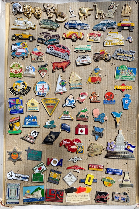 A collection of various vintage pins is displayed on a corrugated cardboard backdrop. The pins feature a variety of designs including cars, animals, flags, logos, and symbols. The pins are colorful and arranged in a somewhat random order, covering most of the cardboard surface.