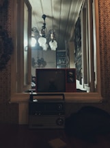A vintage setting with a red old-fashioned television on a window ledge, below which sits a classic radio. The window is open, showing a well-lit interior featuring ornate ceiling lights and a wall lined with picture frames. The ambiance is nostalgic and warm.