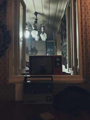 A vintage setting with a red old-fashioned television on a window ledge, below which sits a classic radio. The window is open, showing a well-lit interior featuring ornate ceiling lights and a wall lined with picture frames. The ambiance is nostalgic and warm.