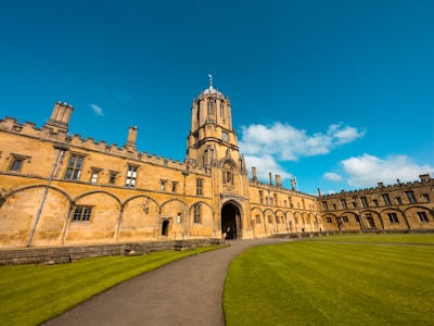 A historic, stone university building with arched windows and detailed architectural elements stands prominently. A tall clock tower rises in the center, flanked by symmetrical wings. A well-maintained lawn and pathway lead up to the building under a clear blue sky with scattered clouds.