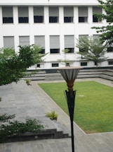 A courtyard with neatly manicured grass, surrounded by a stone pathway and steps. A modern lamp post is central in the foreground. Trees and small potted plants are arranged around the area. In the background, a white building with multiple windows, covered with horizontal blinds, provides a backdrop to the courtyard.