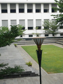 A courtyard with neatly manicured grass, surrounded by a stone pathway and steps. A modern lamp post is central in the foreground. Trees and small potted plants are arranged around the area. In the background, a white building with multiple windows, covered with horizontal blinds, provides a backdrop to the courtyard.