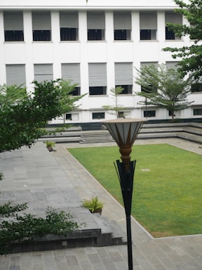 A courtyard with neatly manicured grass, surrounded by a stone pathway and steps. A modern lamp post is central in the foreground. Trees and small potted plants are arranged around the area. In the background, a white building with multiple windows, covered with horizontal blinds, provides a backdrop to the courtyard.