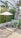 A small outdoor patio setup with a round table and two chairs under a large white patio umbrella. The area is surrounded by lush greenery including palm trees and other plants. A modern building with white walls and decorative grills can be seen in the background.