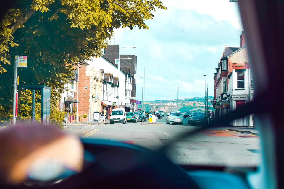 Street scene viewed through car windshield, with buildings and trees visible in foreground