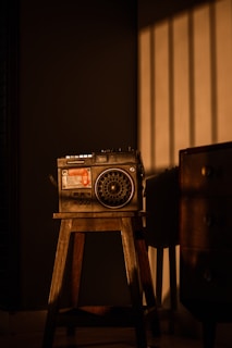 A vintage radio sits atop a wooden stool in a dimly lit room. The warm glow of sunlight casts shadows through vertical blinds, creating an atmospheric play of light and dark on the wall and furniture.