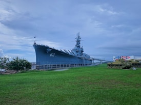 A large battleship with the number 60 is docked on a grassy shore under an overcast sky. To the right, there is a military tank near a building with an American flag painted on it. Tall trees and rocky terrain can be seen in the background.