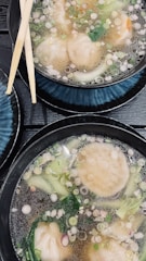 Two bowls of dumpling soup are presented with visible ingredients like dumplings, green vegetables, and a clear broth topped with chopped scallions. Bamboo chopsticks rest on one of the bowls. The setting includes black and blue dishware on a wooden surface, giving a contemporary dining feel.