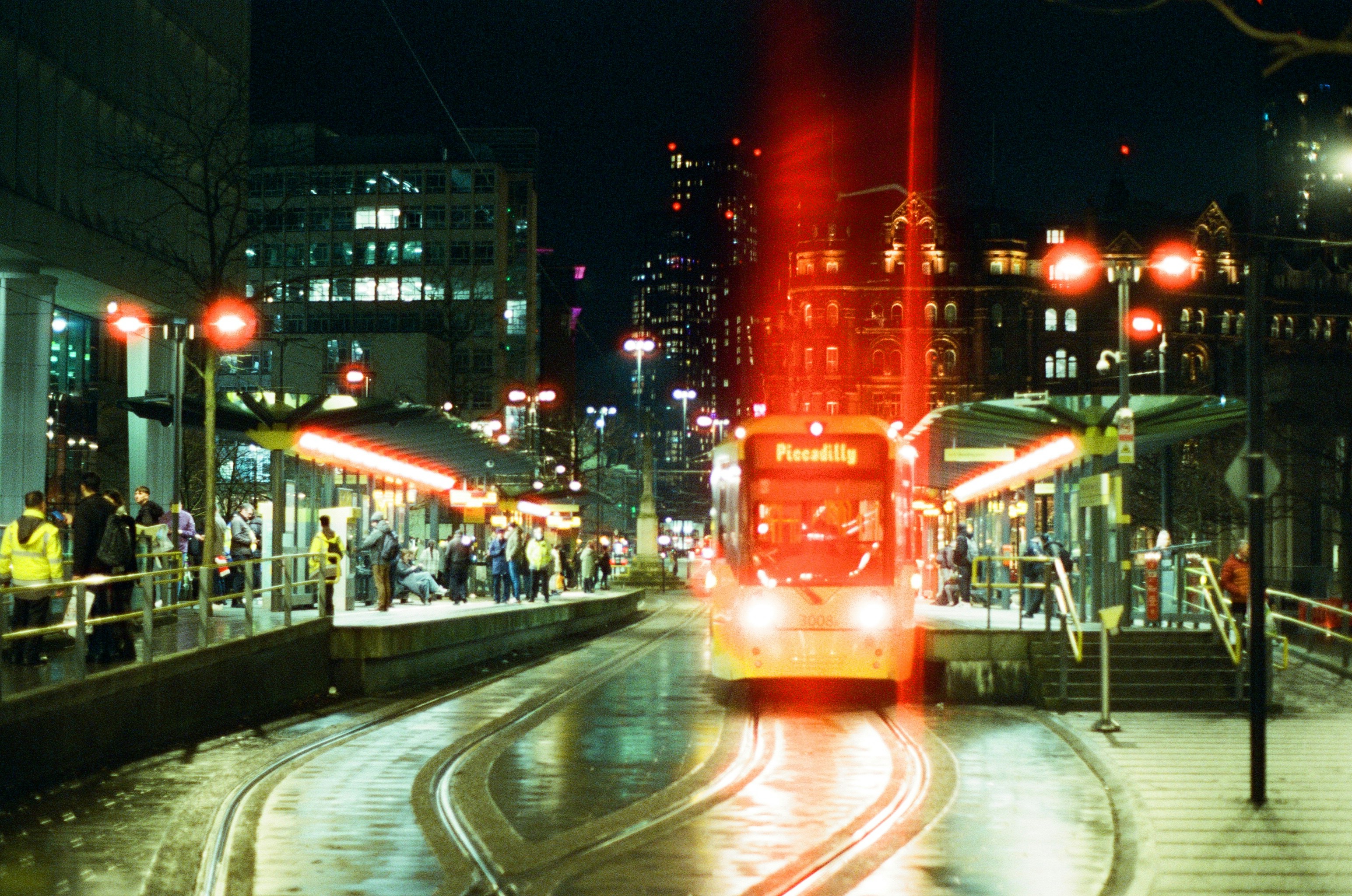 A night-time street scene in Manchester, UK. A large red light-leak covers the tram.