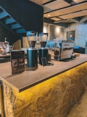 A modern coffee shop interior featuring a coffee bar with an industrial design. Coffee grinders are filled with beans, and there is an espresso machine on the counter. A menu card advertising a 'Big Breakfast' is placed on the countertop. The space has a warm ambiance with natural stone textures and stylish lighting.