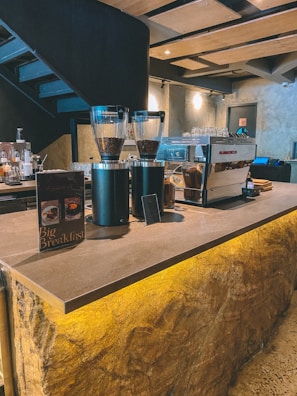 A modern coffee shop interior featuring a coffee bar with an industrial design. Coffee grinders are filled with beans, and there is an espresso machine on the counter. A menu card advertising a 'Big Breakfast' is placed on the countertop. The space has a warm ambiance with natural stone textures and stylish lighting.