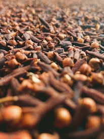 A close-up view of a large pile of cloves showcases their brown, textured surface. The image is sharply focused on the cloves in the foreground, with the depth of field gradually blurring the background.