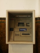 An ATM machine embedded in a wall, featuring a screen, card slot, keypad, and cash dispenser. The surrounding surface is tiled and the machine has a 'ATMANDIRI' logo displayed prominently.