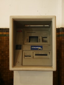 An ATM machine embedded in a wall, featuring a screen, card slot, keypad, and cash dispenser. The surrounding surface is tiled and the machine has a 'ATMANDIRI' logo displayed prominently.