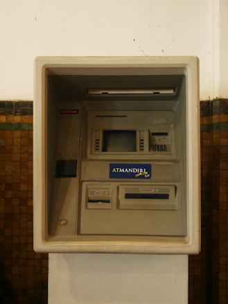 An ATM machine embedded in a wall, featuring a screen, card slot, keypad, and cash dispenser. The surrounding surface is tiled and the machine has a 'ATMANDIRI' logo displayed prominently.