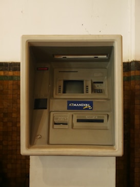 An ATM machine embedded in a wall, featuring a screen, card slot, keypad, and cash dispenser. The surrounding surface is tiled and the machine has a 'ATMANDIRI' logo displayed prominently.