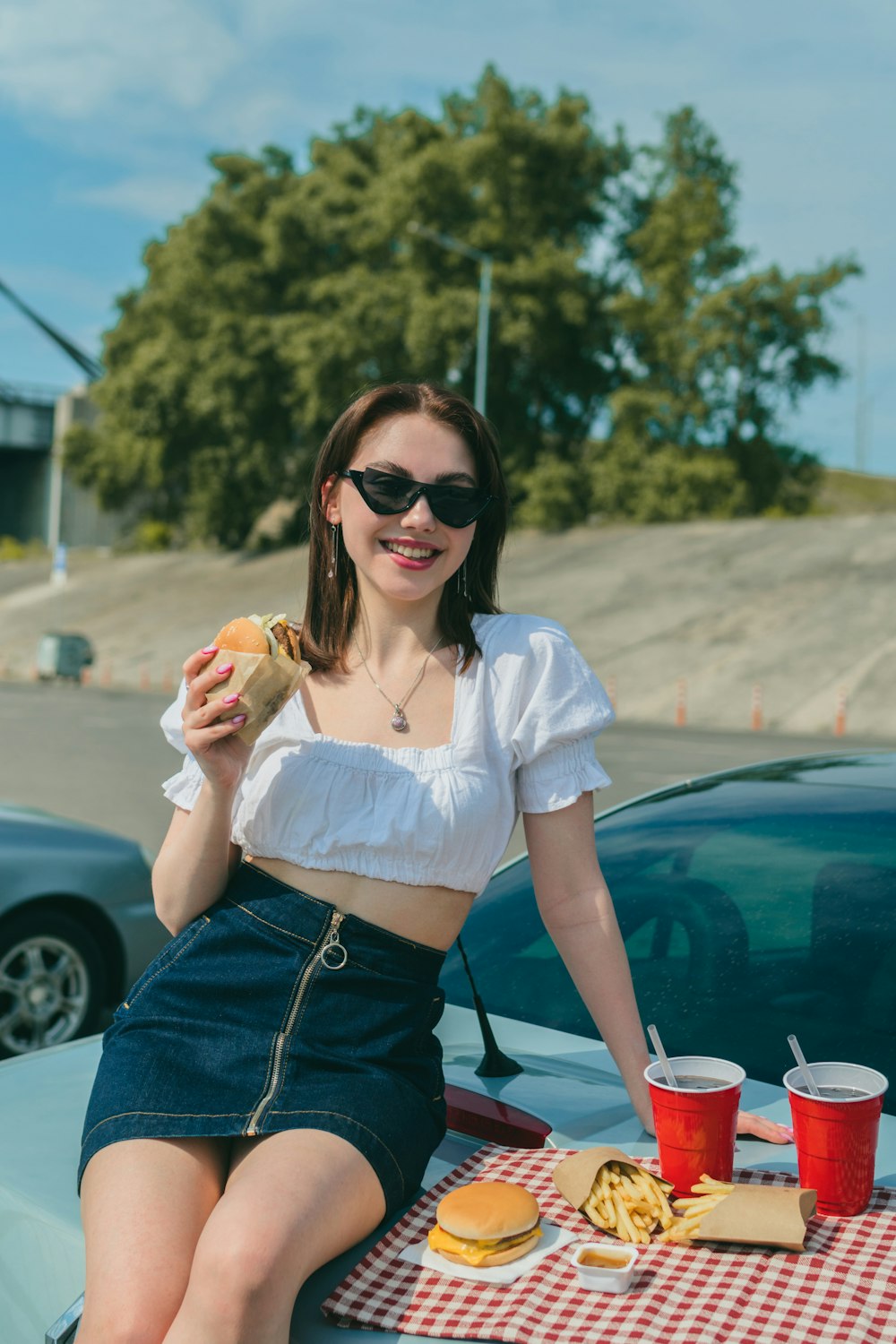 a woman sitting on the hood of a car eating a hamburger and fries