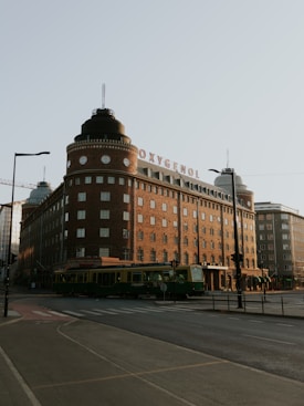 A large, historic brick building with a sign that reads 'OXYGENOL' on the roof. The building features rounded corners and numerous windows. A green tram is passing by in the foreground on a city street. The sky is bright and clear, suggesting a sunny day.
