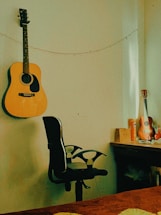An acoustic guitar hangs on the wall next to a black office chair. A string of lights is draped across the wall. A desk is positioned in the corner with a table lamp and a ukulele placed on it alongside various small items and papers.