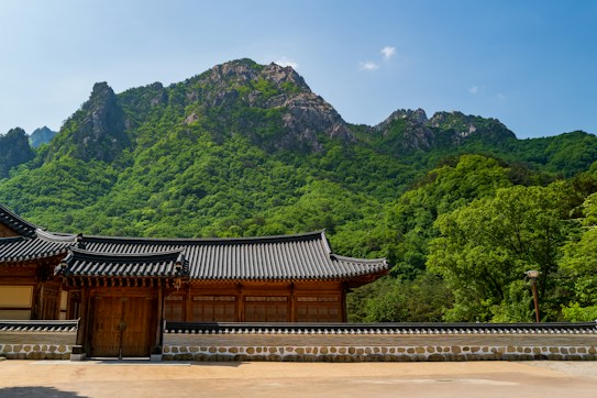 A traditional Korean building with a wooden gate and tiled roof sits in front of a lush, green mountainous landscape. The sky above is clear and bright blue, complementing the vibrant greenery of the dense forest on the mountainside. Architectural elements include intricate woodwork and classic stone walls.