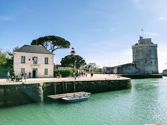 A picturesque waterfront scene featuring a stone building with flags, a circular stone tower, and a lighthouse in the distance. Several people are walking along the promenade, enjoying a sunny day. A small boat is docked in the calm, turquoise water. The sky is clear and blue, adding to the tranquil atmosphere.