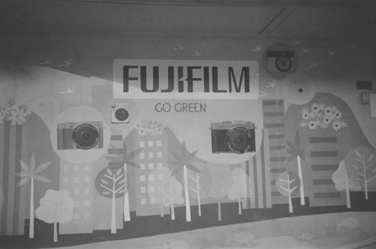A mural featuring the brand name Fujifilm prominently at the top, with the slogan 'Go Green' displayed underneath. The artwork includes illustrations of cameras, trees, buildings, flowers, and birds, suggesting a blend of technology and nature.