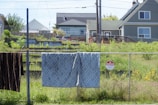 A residential area with two houses in the background. In the foreground, there are items hanging on a chain-link fence, including a patterned blanket and a dark-colored fabric. A 'No Trespassing' sign is attached to the fence. The scene includes a grassy yard with some overgrown plants.