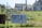 A residential area with two houses in the background. In the foreground, there are items hanging on a chain-link fence, including a patterned blanket and a dark-colored fabric. A 'No Trespassing' sign is attached to the fence. The scene includes a grassy yard with some overgrown plants.
